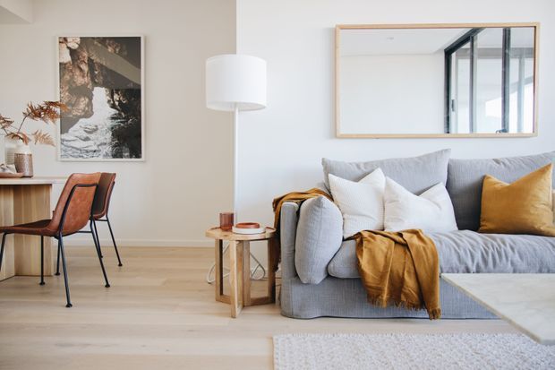Bright, softly styled living room with a grey upholstered sofa, white and ochre cushions, a timber side table, floor lamp, pale timber flooring, and a dining/work table with leather chairs. A large wall mirror reflects black-framed glazing, while neutral walls and restrained decor create an airy contemporary setting.