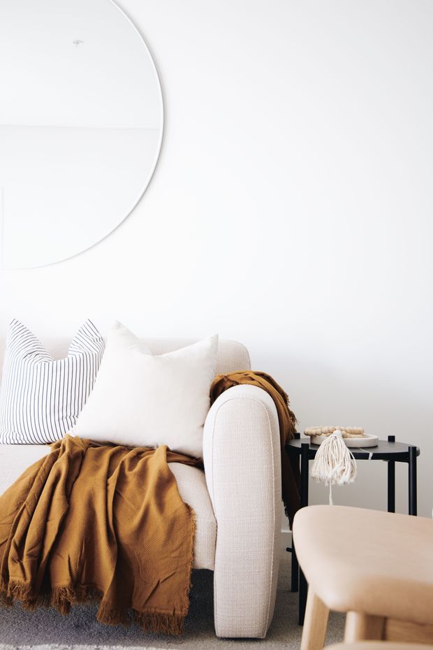 Bright, minimalist living room vignette featuring a cream upholstered armchair, white and striped cushions, and a mustard-brown textured throw. A black side table with small decor sits beside the chair, with a pale wood coffee table in the foreground and a large round mirror on the white wall.
