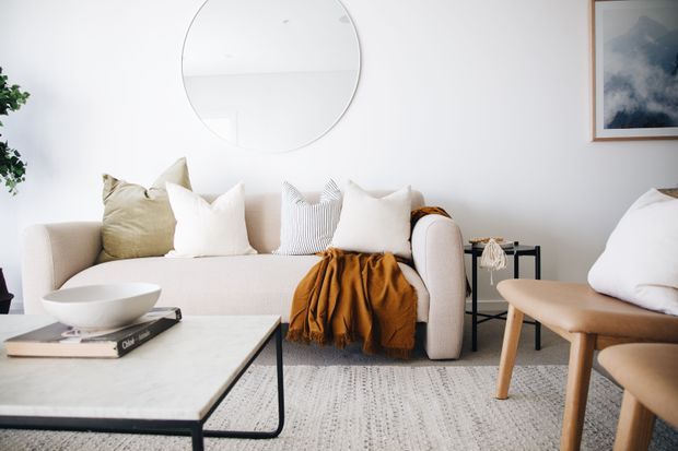 Bright, serene contemporary living room with a cream upholstered sofa layered with neutral cushions and a mustard throw, a pale stone or marble-look coffee table, light textured rug, black side table, and a timber-framed armchair. White walls feature a large round mirror and framed artwork.
