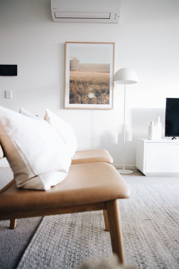 Bright, softly styled living room with a tan leather or suede lounge chair in the foreground, white cushions, a pale textured rug, a white media console and television, a slim white floor lamp, framed landscape artwork, and a wall-mounted air conditioner. The room has a clean, neutral and understated contemporary aesthetic with abundant natural light.