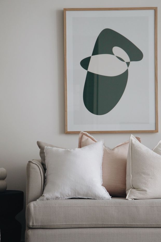 A close-up of a softly styled living room featuring a beige upholstered sofa with layered white, cream, and pale pink cushions. A framed abstract artwork with a natural timber frame hangs above the sofa against a light neutral wall. A dark side table and sculptural neutral-toned decor are partially visible at the left edge. The overall look is calm, refined, and contemporary with warm neutral tones.