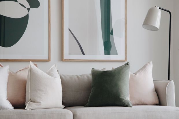 Close-up of a softly styled living room seating area featuring a light neutral upholstered sofa with layered cream, blush, beige, and deep green cushions. Two abstract framed artworks hang above the sofa, accompanied by a slim black floor lamp with a white shade. The room has a calm, refined, contemporary appearance with a muted natural palette.