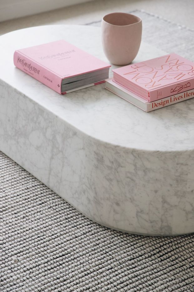 Close-up interior detail of a low, rounded rectangular coffee table with a white veined marble finish, styled with pink design books and a blush-toned ceramic cup. The table sits on a textured grey and off-white woven rug in a softly lit, minimalist living space.