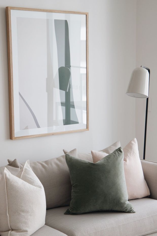 A close-up of a softly styled living room featuring a light neutral upholstered sofa with cream and blush cushions, a deep sage-green accent cushion, a minimalist abstract artwork in a light timber frame, and a slender black floor lamp. The space has a calm, refined, contemporary aesthetic with a muted neutral palette and natural timber detailing.