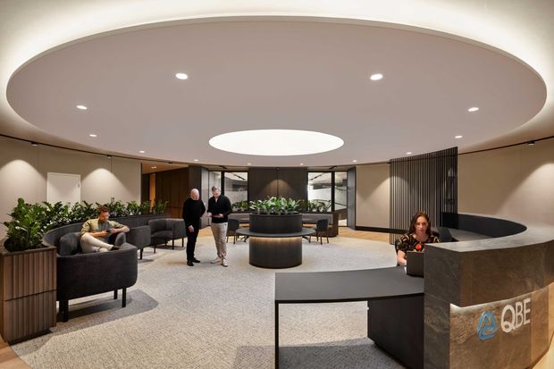 A sophisticated commercial reception and lounge area with a large curved oval ceiling feature, central circular skylight or illuminated ceiling opening, recessed downlights, a curved reception desk, and a dark slatted privacy screen. Neutral taupe walls, carpeted flooring, black and charcoal furniture, large planters, and integrated greenery create a refined contemporary office lobby. Several people are present, and the reception desk bears a visible QBE logo.