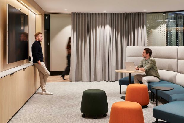 Contemporary interior lounge or collaboration area with built-in timber cabinetry and wall-mounted display on the left, floor-to-ceiling grey curtains, upholstered banquette seating, a small table with a laptop, and colourful rounded ottomans. The space appears to be a refined commercial or workplace breakout area with carpeted flooring and glazed partitions.