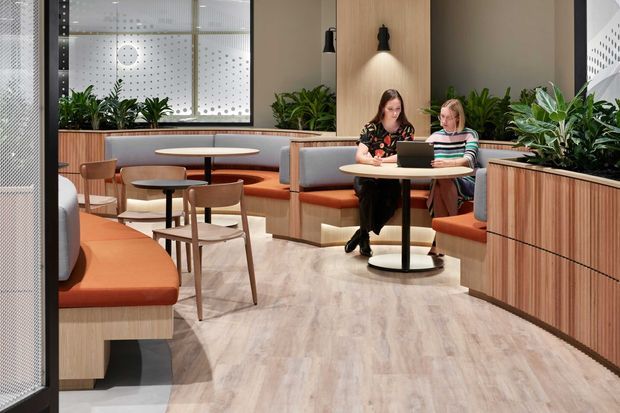 A bright contemporary commercial lounge or hospitality-style breakout area with built-in banquette seating, small round café tables, timber-look vertical slat joinery, integrated planter boxes, and frosted patterned glazing. Warm terracotta upholstery contrasts with pale timber flooring and neutral walls. Two people are seated at a table, while pendant and wall lighting create a welcoming workspace atmosphere.