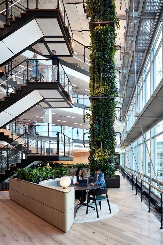 A bright multi-level commercial interior with dramatic zigzag staircases, glass balustrades, extensive floor-to-ceiling glazing, and a tall vertical planted feature column. A small collaborative workspace with built-in timber planter joinery, a round table, chairs, and two people working on laptops occupies the foreground. The space has an open atrium-like volume, pale timber flooring, exposed dark stair framing, and a patterned geometric ceiling.