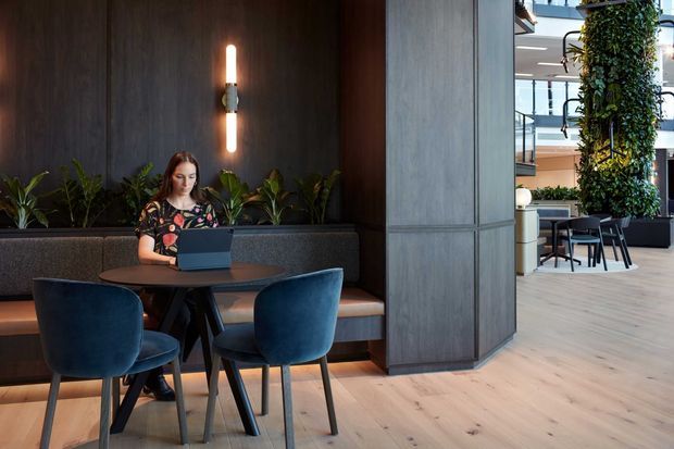 A contemporary commercial interior showing a woman working on a laptop at a small round table in a dark upholstered booth. Deep blue chairs, warm wood flooring, dark timber wall panelling, integrated planter boxes, and a slim vertical wall light create a refined hospitality or workplace lounge setting. An open seating area with additional tables, chairs, and a tall indoor planting feature is visible beyond the central partition.