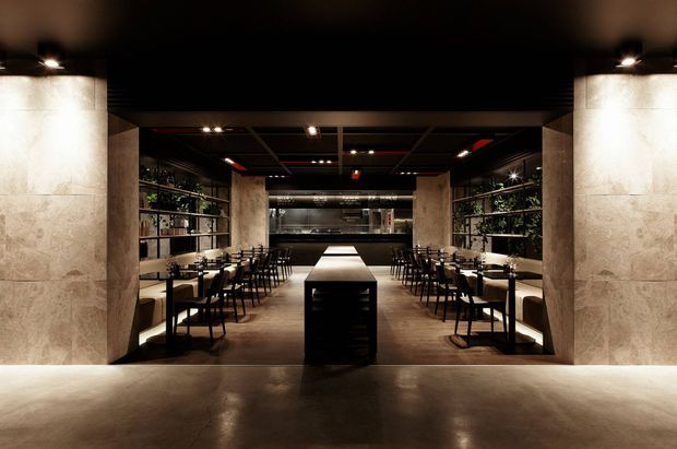 A dark, sophisticated commercial dining space viewed from the entrance, with rows of small tables and chairs on both sides, a central island or service counter, open shelving with greenery and objects, and a rear bar or service area. Large pale stone-clad columns and walls contrast with the black ceiling, dark metal shelving, warm concealed lighting, and polished dark flooring. The overall atmosphere is intimate, dramatic, and refined.