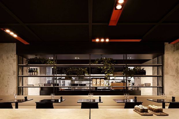 A dark, sophisticated commercial dining space viewed across a pale stone counter. The room features black metal open shelving with plants, bottles, crockery and serving items, a textured pale stone or concrete wall finish, black tables and chairs, and warm linear ceiling lighting with orange-red accents. The composition suggests a contemporary restaurant or café interior with a refined industrial character.