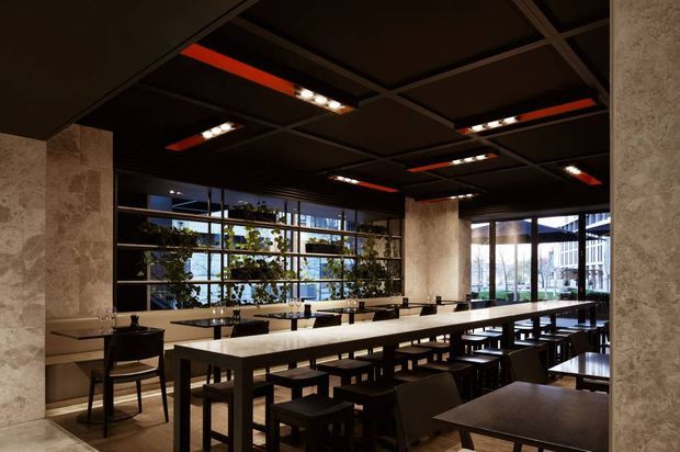 A dark, sophisticated communal dining or hospitality interior with long marble-topped tables, black stools and chairs, full-height glazing, and integrated planter shelving. The ceiling features a dramatic black exposed grid with recessed linear lighting and red accent panels, while textured stone-clad columns frame the space. The image appears to show a restaurant, café, or commercial dining area.