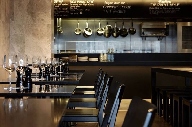 A dark, refined restaurant interior viewed across a long dining counter or communal table. Black and deep brown timber surfaces, aligned dining chairs, wine glasses, place settings, hanging metal cookware, stacked plates, a tiled service backsplash, and a chalkboard menu create a polished commercial hospitality setting.