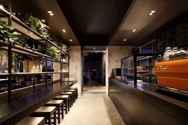 A dark, sophisticated café interior viewed down a central aisle. Open black metal shelving with plants, products and stools lines the left side, while a long dark service counter with a glass pastry display, chalkboard menu and bright orange espresso machine occupies the right. The space features polished concrete flooring, dark ceiling panels, recessed lighting and stone-textured wall surfaces, creating an industrial yet refined atmosphere.