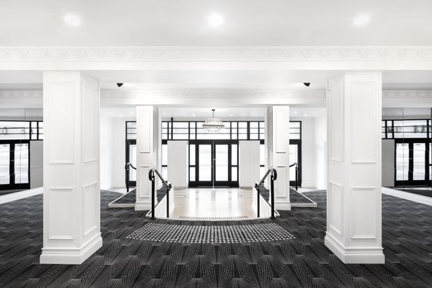 A grand, symmetrical commercial interior lobby or entrance hall with white paneled columns, ornate white ceiling detailing, black-framed glazed doors and windows, a central staircase with metal handrails, and dark patterned carpet surrounding a bright tiled or stone central landing. The space has a refined, high-contrast monochrome appearance and strong geometric composition.