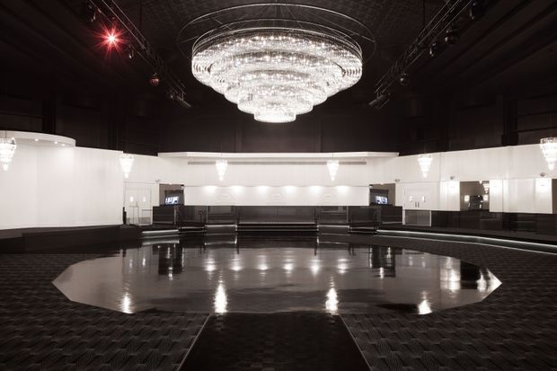 A dramatic, monochromatic interior of a large entertainment or event space. A glossy dark octagonal dance floor or central floor feature is surrounded by black patterned flooring, curved white illuminated walls, mirrored surfaces, and tiered dark seating or bench joinery. A large tiered crystal chandelier dominates the ceiling, with theatrical stage lighting and wall-mounted displays adding to the venue-like atmosphere.