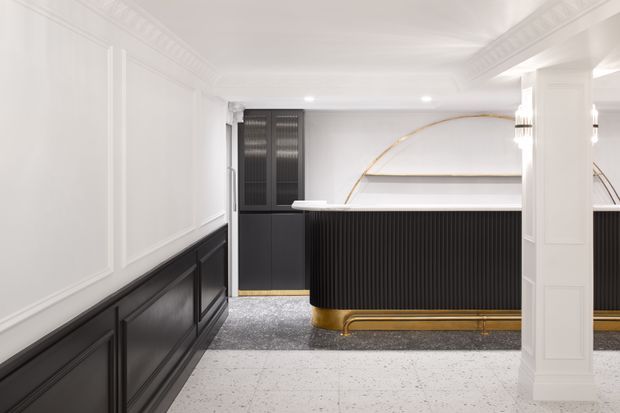 A refined reception or service counter area with strong black-and-white detailing. The space features white decorative wall panelling and ornate ceiling cornices, a black fluted counter front with a white countertop, brass-gold plinth detailing, dark cabinetry, and a pale terrazzo-style floor. Curved brass detailing and illuminated wall features add a luxurious Art Deco character.