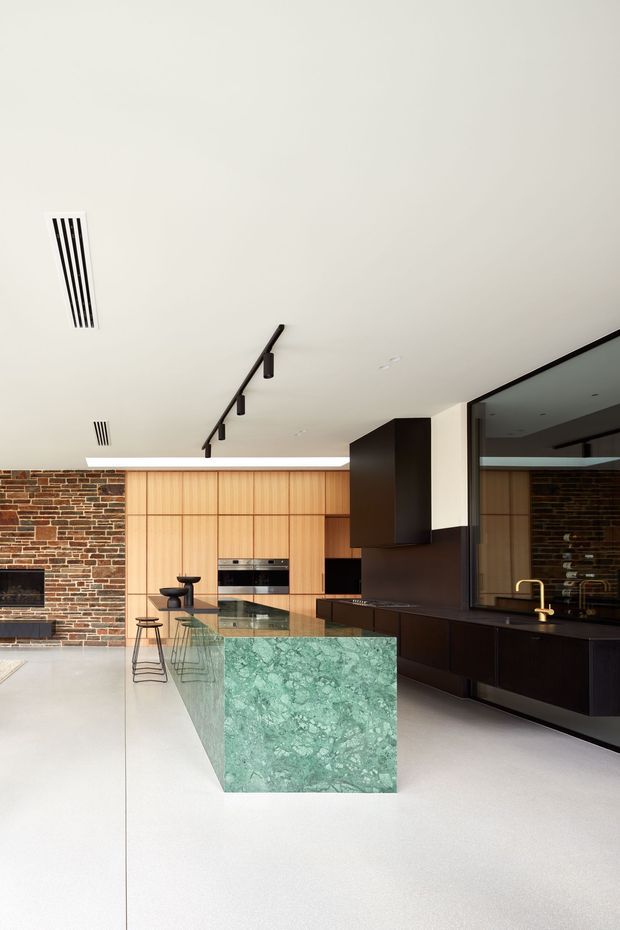 Contemporary open-plan kitchen with a large green stone island, dark built-in cabinetry, timber wall cabinetry, exposed brick feature wall, integrated appliances, and black track lighting. A reflective dark splashback and brass tap add contrast, while the pale floor and ceiling keep the room bright and spacious.