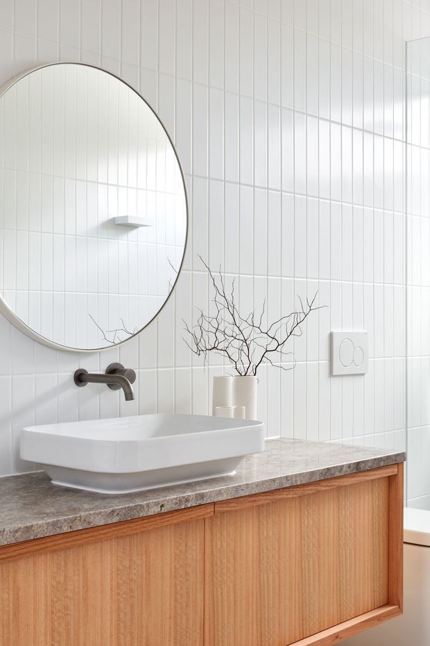 Bright, minimalist bathroom vanity with a large round framed mirror, white vertically lined ceramic wall tiles, a white vessel basin, brushed dark metal wall-mounted tapware, a grey stone-look benchtop, and warm timber cabinetry. A simple branch arrangement adds a natural decorative accent.