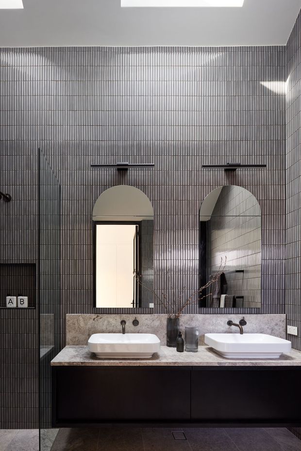 A refined contemporary bathroom with a double vanity, two white vessel basins, dark wall-mounted tapware, and a dark timber or painted vanity cabinet topped with a grey stone benchtop. The walls are clad in narrow vertically ribbed grey tiles, complemented by two arched mirrors and arched wall openings. A frameless glass shower enclosure is visible on the left, with dark tiled flooring and skylights providing natural light.