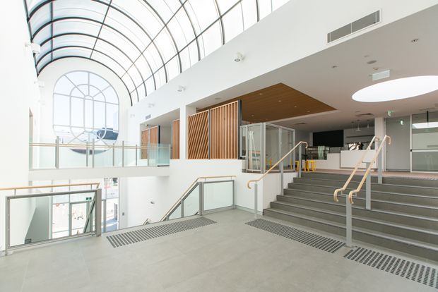 Bright contemporary multi-level interior atrium with a sweeping glazed barrel-vault roof, tall arched glazing, glass balustrades, broad staircases, pale tiled or concrete flooring, and warm timber slatted wall and ceiling features. The open circulation space includes accessible handrails, recessed lighting, and a mezzanine level, suggesting a public or commercial building.