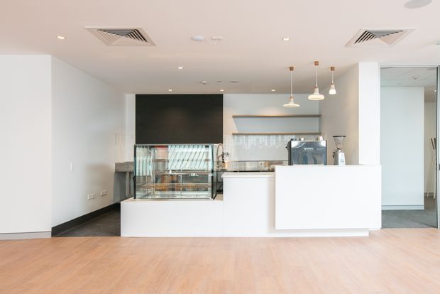 Bright contemporary commercial café or food-service interior with a compact white service counter, glass display case, coffee equipment, tiled splashback, pendant lights, recessed ceiling lighting and pale timber-look flooring. The space is mostly white and minimal, with a dark feature wall behind the counter and an open doorway to a secondary room.