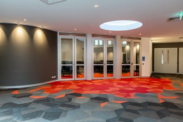 Interior view of a contemporary commercial lobby or circulation area. The space has dark curved walls with recessed spot lighting, a vivid geometric patterned carpet in red, orange, pink, grey and charcoal, glazed meeting-room doors with visible tables and chairs beyond, double access doors, and a large oval recessed ceiling light.