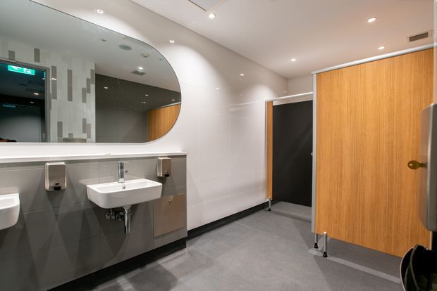 A clean contemporary commercial bathroom interior with multiple wall-mounted basins, a large rounded-corner mirror, tiled walls, grey flooring, stainless-steel dispensers, and timber-faced restroom cubicle doors. Recessed ceiling lights and a dark accent wall create a functional, polished public washroom setting.