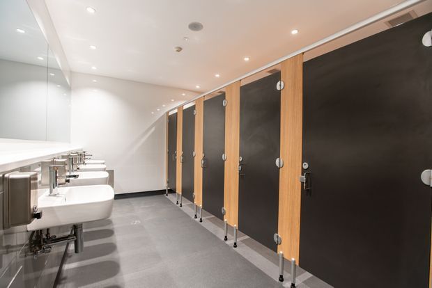 A clean, modern public restroom with a row of white wall-mounted basins and chrome faucets along the left wall, large frameless mirrors above, and dark charcoal partition cubicles with warm timber-coloured vertical panels and exposed support feet. The space has a smooth white ceiling with recessed downlights and a grey tiled floor.