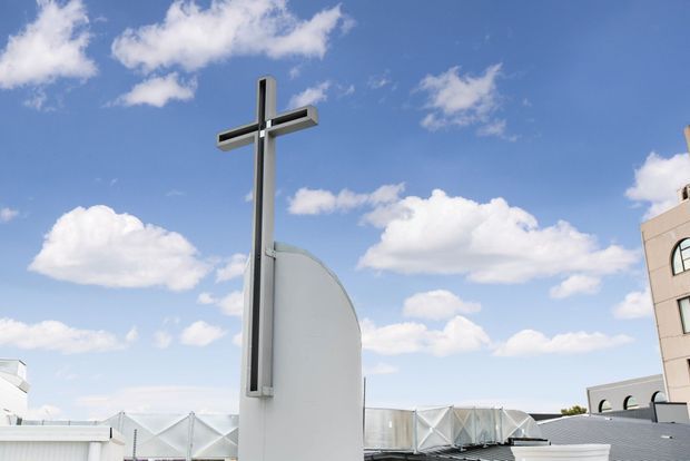 Exterior architectural detail of a modern church or religious building, featuring a tall minimalist metal cross mounted on a sculptural white curved wall. The background includes a bright blue sky with clouds and portions of a contemporary building complex with geometric roof structures.