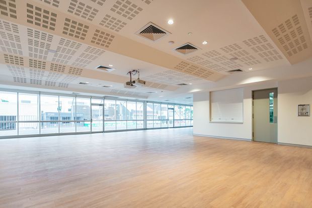 A large, bright open-plan commercial interior with extensive floor-to-ceiling glazing along one side, overlooking an urban setting. The room has a warm timber-look floor, pale walls, a suspended acoustic perforated ceiling with recessed lighting and exposed HVAC grilles, and a ceiling-mounted projector. A whiteboard and internal door are visible on the right, suggesting a meeting, training, or classroom space.