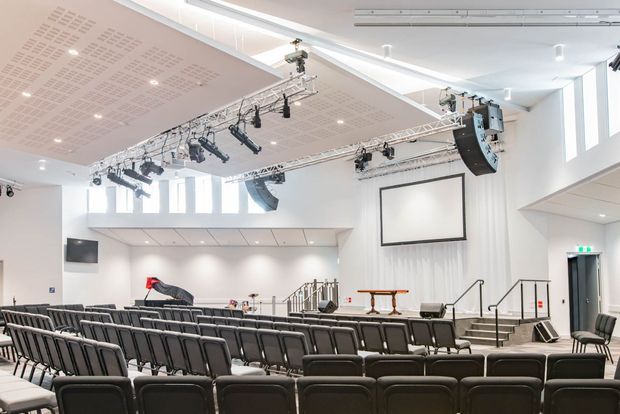 Bright contemporary auditorium or lecture theatre viewed from the rear seating area. Multiple rows of upholstered fixed seats face a raised stage with a projection screen, lectern or table, stairs, handrails and extensive suspended theatrical lighting and audio equipment. The room has white walls, high ceilings, clerestory-style windows, acoustic ceiling panels and a clean institutional or performance-venue fit-out.
