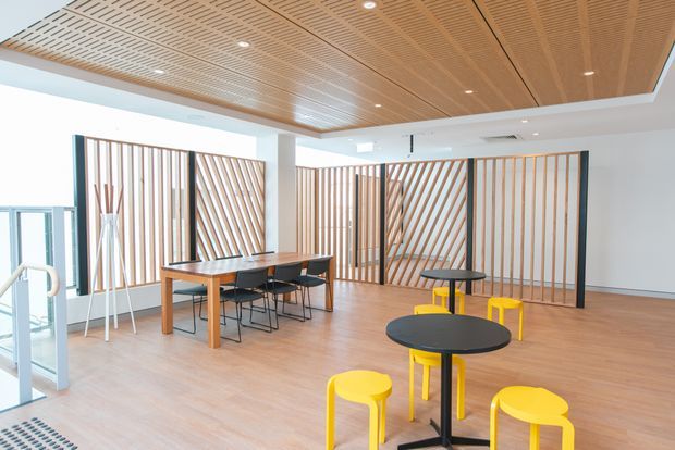 Bright contemporary communal interior with an open-plan dining and breakout area. The space features a warm timber slatted ceiling, light timber flooring, white walls, recessed downlights, and multiple timber-and-black framed decorative screening panels. A long timber dining table with black chairs occupies the left side, while round black tables with vivid yellow stools create informal seating in the foreground. Glazing and a glass door are visible at the left.