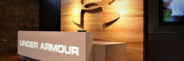 A dark, modern commercial reception area featuring a white Under Armour branded desk in front of a large illuminated timber feature wall with the carved Under Armour logo. Black ribbed wall panels and a partial framed display are visible at the sides.