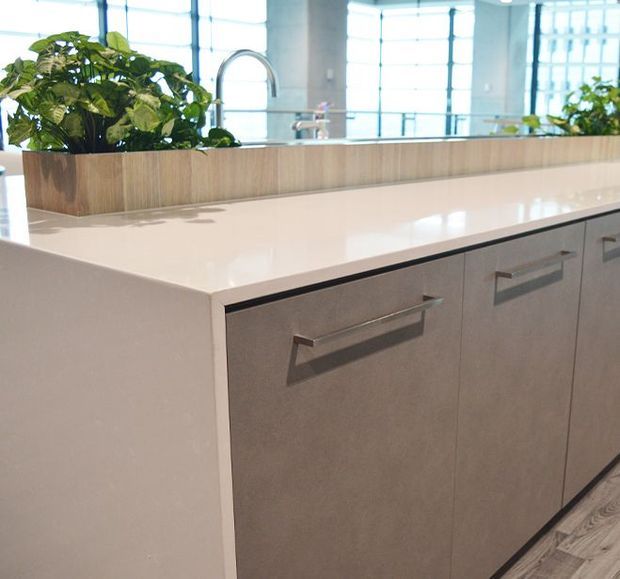 Close-up of a contemporary kitchen island with a bright white solid-surface benchtop, warm wood planter box, integrated greenery, and grey flat-panel cabinetry with long metal handles. Large glazed windows and concrete structural columns are visible in the background.