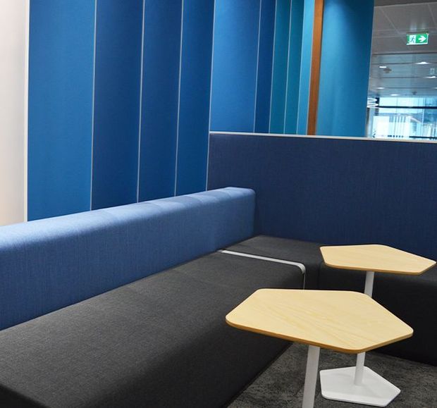 Contemporary commercial lounge or waiting area with deep blue upholstered banquette seating, charcoal fabric seating, light timber-topped side tables, and blue vertical wall panelling. A glazed office or corridor is visible beyond the partition, reinforcing the workplace setting.