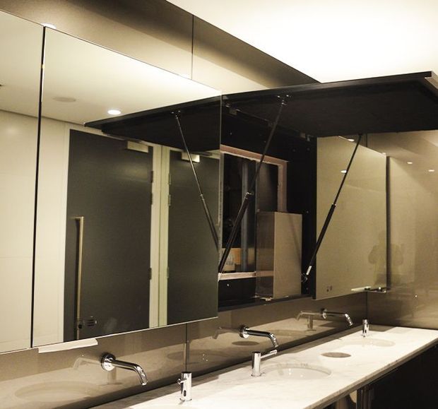 A contemporary commercial-style bathroom vanity with a long white counter, multiple integrated basins and chrome wall-mounted faucets. Large mirrored wall panels reflect the dark doors and vanity area, while black overhead mirrored cabinet doors are propped open with gas struts. Recessed ceiling lighting and neutral beige-grey finishes create a clean, functional atmosphere.