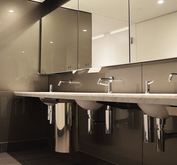A contemporary commercial bathroom or washroom with a long pale stone vanity, multiple integrated white basins, chrome wall-mounted taps, mirrored upper wall panels, dark glossy wall and floor tiles, and exposed polished metal plumbing. Recessed ceiling lights provide even illumination.