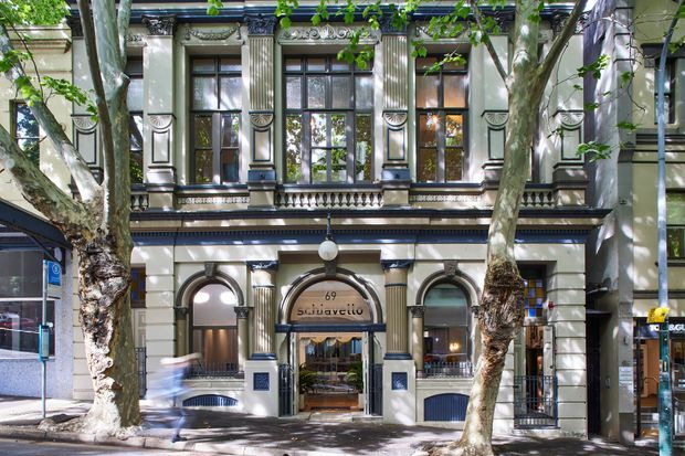 Street-level photograph of an ornate historic multi-storey commercial building facade, framed by mature street trees. The cream-painted masonry frontage features strong classical detailing, fluted pilasters and columns, arched openings, decorative balustrades, dark trim, tall multi-pane windows, and a central glazed entrance with signage reading “Silk...”.