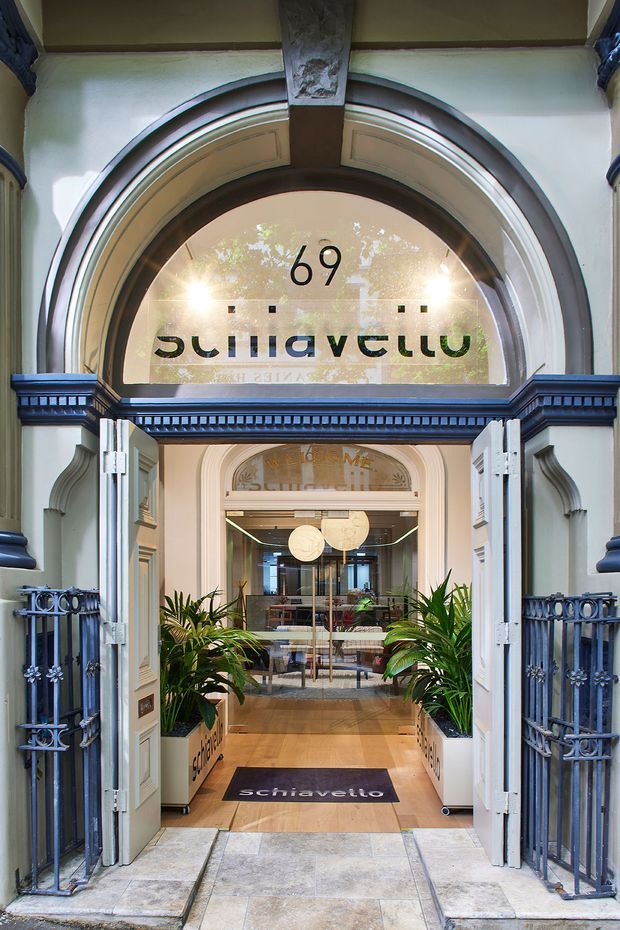 Front-facing view of an ornate commercial building entrance with open double doors, a large arched fanlight bearing the number 69 and mirrored Schiavello signage, decorative blue-painted mouldings, wrought-iron gates, stone paving, and potted plants. The entry opens into a refined modern showroom or office reception with glass partitions, timber flooring, contemporary lighting, and seating visible inside.