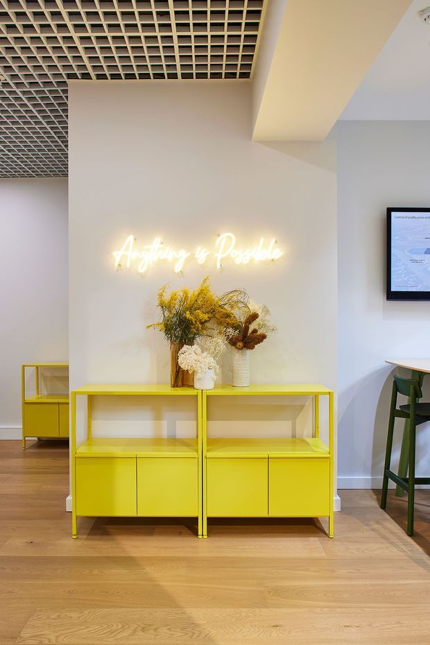 A bright contemporary interior reception or display area with vivid yellow built-in-style display cabinets against a white wall. A glowing cursive neon sign reading “Anything is Possible” hangs above decorative dried floral arrangements. The space has warm timber flooring, a distinctive open grid ceiling, and a partial high table with green stools visible at right.