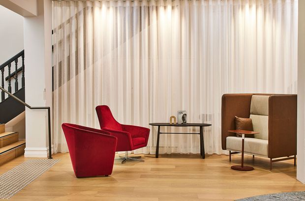 A refined living room with warm timber flooring, full-height sheer curtains, and a dramatic red lounge chair paired with a sculptural red armchair. A high-backed upholstered occasional chair, slim round coffee table, framed artwork, and a staircase with dark balustrade complete the space. The room has a calm, gallery-like atmosphere with strong mid-century-inspired furnishings.