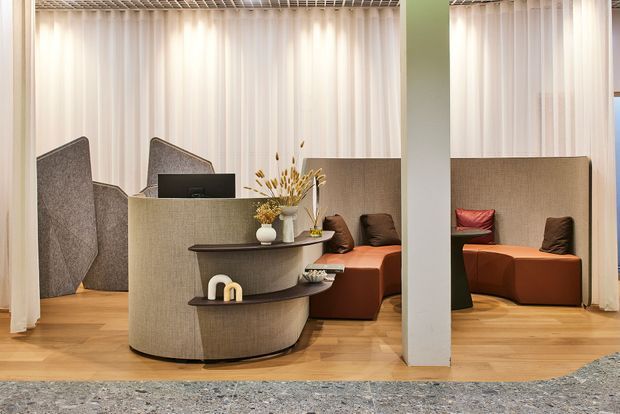 A contemporary commercial lounge or reception area with a curved upholstered reception desk, integrated display shelves, computer workstation, partitioned seating booths, warm timber flooring, floor-to-ceiling sheer curtains, and a pale structural column. Rust and brown upholstered seating adds warmth, while a patterned grey carpet occupies the foreground.