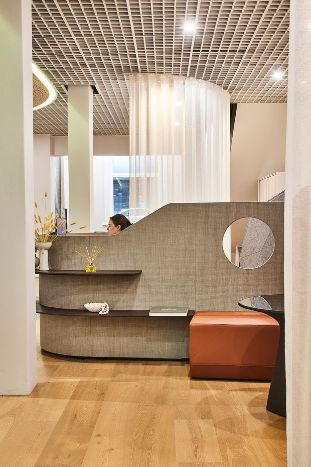 A refined contemporary commercial interior, likely a reception or showroom space, viewed through a wide doorway. The room features a curved textured taupe reception counter with integrated dark shelving, a circular wall mirror, an orange upholstered ottoman, light timber flooring, pale walls, and a dramatic suspended translucent curtain-like cylindrical feature. A white geometric grid ceiling with recessed lighting adds a distinctive architectural element.