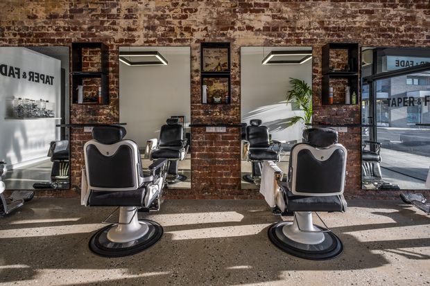 A bright industrial-style barbershop interior with multiple vintage barber chairs arranged in front of large mirrors. Exposed aged brick walls, dark timber shelving, framed artwork, concrete-look flooring, and strong sunlight create a characterful commercial grooming space. Street frontage and neighbouring buildings are visible through the glazing.