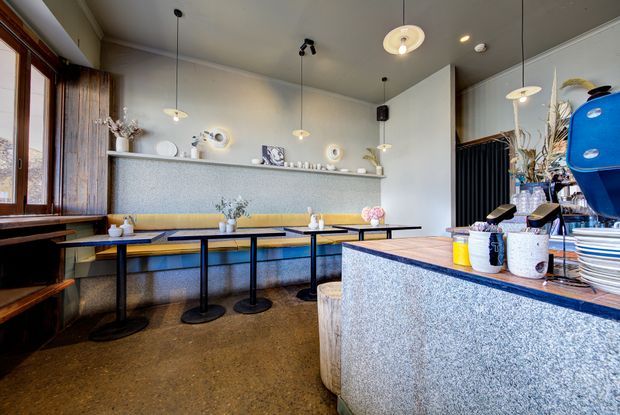 A stylish café interior with a long upholstered banquette, compact dark-framed tables, a textured stone or terrazzo service counter, timber-framed windows, pendant lighting, and curated tabletop and wall décor. The space has a warm mid-century-inspired atmosphere with blue, yellow, grey, and timber accents.