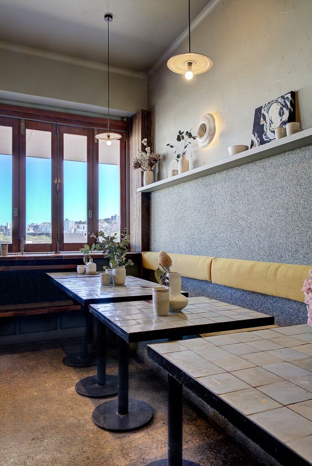 A warmly styled cafe dining area with tiled tables, black pedestal bases, a long upholstered banquette, textured walls, timber-framed windows, pendant lighting, and decorative shelving. Urban views are visible through the windows.