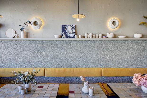 A stylised contemporary hospitality or dining interior featuring a long speckled grey counter with an upholstered mustard-yellow banquette below. The textured off-white wall behind the counter displays ceramic vessels, sculptural circular wall lights, artwork, and decorative objects. Low tiled tables with warm timber and dark inset details sit in front, accompanied by floral arrangements and tableware.