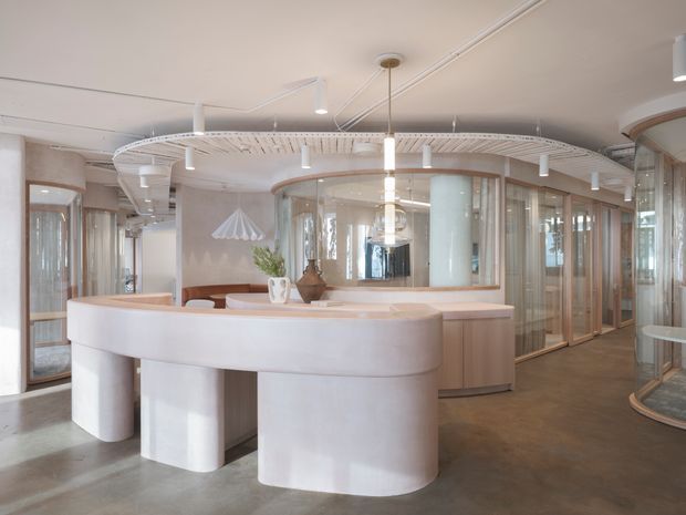 A bright, refined commercial interior with a large curved white reception or service counter in the foreground. The space features rounded built-in forms, warm timber cabinetry and framing, extensive curved glass partitions, a polished concrete floor, suspended linear lighting, and a distinctive white slatted or ribbed ceiling feature. The open-plan layout appears contemporary and softly minimalist.