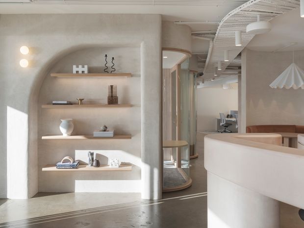 A bright, softly sculpted contemporary commercial interior with rounded plaster walls and a built-in arched shelving niche displaying small objects and books. A curved white reception or service counter occupies the foreground, while a workstation and rounded glass-fronted display or meeting elements extend into the background. Exposed white cable trays and services run across the ceiling, contrasting with the refined, warm neutral finishes. Pendant lighting, recessed wall lights, timber shelving, and polished concrete flooring complete the space.